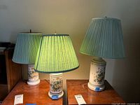 Three ceramic table lamps arranged on a wooden surface, lit from one lamp. Each has a white ceramic base with floral design and light green pleated shades.