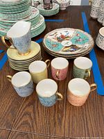 Photo of six demitasse cups in pastel colors with gold handles and rims, arranged near matching saucers with a creamer on top.
