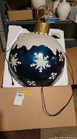 Full view of the large blue and white snowflake Christmas ornament ball with power cord and packaging box visible underneath.