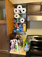Kitchen counter surface with various cleaning products grouped together including sprays, bottles, and rolls of paper towels visible beside a stove and cabinet.