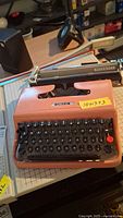 Full view of orange Olivetti Lettera 22 manual typewriter with black keys on a wooden table with various items in background.