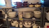Shelf holding white ceramic bowls, stacked plates, white ceramic mugs and cups, two white lidded casserole dishes, brown glazed ceramic lidded serving dishes, and beige crocks.