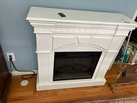 White electric fireplace heater with mantel and decorative molding, plugged in at wall outlet.
