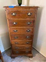 Front view of medium wood-tone seven drawer chest with brass handles on each drawer showing overall condition and size relative to a carpeted floor.