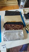 Five evening bags arranged on table, showing black fabric bag with bow, gold clutch, velvet patterned clutch, silver sequined clutch, and black satin-like clutch.
