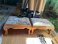 Pair of carved wood foot stools with floral needlepoint upholstered tops shown on wood floor near window.