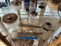 Three pieces of cut glassware with silver metal lids displayed on glass shelf.