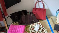 Photo showing four handbags on a table including red leather, dark brown suede, pink fabric, and a white floral fabric with green rope handles bag