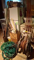 View of three rolled green garden hoses coiled on the floor, a white hose rack with an attached brown hose, several garden tools stored in a cardboard box, and various stakes and trellises leaning against a wall.
