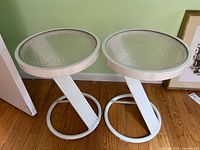 Two round white patio side tables with glass tops placed on wooden floor against a green wall.