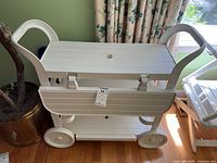 White plastic tea cart with two dropleaf side panels folded down, four wheels, and curved handles, shown from the side with floor and plant background.