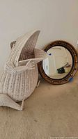 Photo showing the frog-shaped wicker basket sitting next to the round wooden framed mirror on a carpeted floor. Basket has an open mouth design and is made of white wicker.