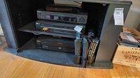 Wide view of black Sony Blu-ray/DVD player, VCR, DVD/CD changer stacked on black shelf with multiple remotes in holder.
