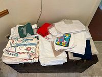 Two piles of vintage linens and cloths including embroidered tablecloths, towels, and vintage baby clothes on top of a wooden chest against a wall.