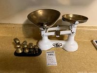 Full view of the vintage white and brass balance scale with the set of brass weights on a black tray and a note showing lot number and details.
