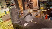 Two aluminum watering cans on a table, one with a long spout and rose nozzle, the other a smaller unnamed can.