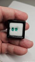Pair of vivid green oval cut Columbian emerald gemstones displayed in black jewelry box with white cushion.