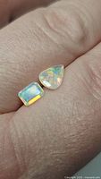 Two loose opal gemstones resting on a finger to show size and color. One is a pear cut, the other emerald cut. Both show vibrant multi-colored flashes.