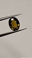 Close-up image of oval cut tourmaline gemstone held by tweezers, showing its deep green-yellow color and clarity.