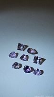 Nine natural amethyst gemstones varying in color from light pink to purple with mixed faceted cuts including round, oval, triangular and square shapes arranged on a plain background.