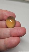 Photo of a hand holding a large oval cabochon lemon citrine gemstone showing smooth polished surface, lemon yellow color, and size relative to fingers.