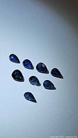 Nine natural pear cut blue sapphire gemstones with deep blue color and VS clarity, laid out on a plain white surface under bright lighting.