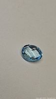 Close-up photo of the single oval blue topaz gemstone on a neutral background showing the checkerboard cut facets and color