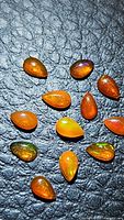Multiple pear cabochon fire opal gemstones displayed on a textured black surface showing their deep orange color and flashes of green.
