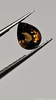 Close-up photo of a pear-shaped multicolored orange yellow tourmaline gemstone held by tweezers against a neutral background showing the gem's rich fire and clarity.