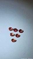 Six natural Padparadcha sapphires arranged on a flat surface showing their pink to orange-pink color and faceted diamond cut.