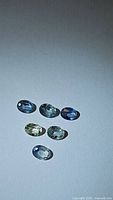 Six oval cut natural sapphires in pastel shades arranged on flat surface under light showing various subtle color hues and clarity.
