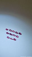 Top-down view of eleven oval cut pinkish-red rubies arranged in rows on a plain light surface, showing clarity and color variation