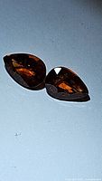 Pair of two pear-shaped untreated Spessartite garnet gemstones with deep vibrant orange color, placed on light blue background.