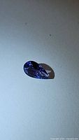 Deep blue-purple pear-shaped tanzanite gemstone with facets visible, placed on a light-colored background.