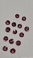 Close-up of 13 small round cut sapphires on a white background showing pink, purple, and orange hues, confirming lot contents and condition.