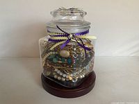 Full view of glass jar filled with jewelry, lid on, ribbons visible, on wood base