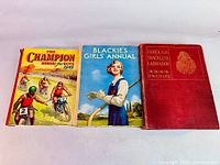 Three antique hardcover books placed side by side, showing front covers of The Champion Annual for Boys 1943, Blackie's Girls Annual, and Through Trackless Labrador by H. Hesketh Prichard