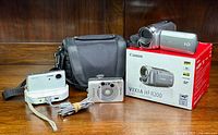 Lot contents showing the Canon Vixia HF R200 camcorder on its box, Canon ELPH Jr. compact camera, Sony Cybershot on docking station, and carrying bag.