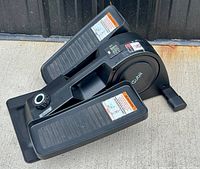 Side view of black Cubii Jr. under desk elliptical showing foot pedals with warning labels, adjustable resistance knob, and brand logo.
