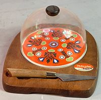 Wooden cheese board with colorful orange floral patterned domed cover and matching cutter resting in a carved slot.