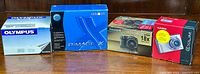 Four boxed compact digital cameras: Olympus (white/blue box), Minolta DiMAGE X (blue/black box), Fujifilm FinePix S5600 (black/gold box), Casio Exilim EX-Z60 (red/white box).