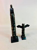 Two black resin Northwest Coast style totem pole sculptures, one tall and one shorter with broad wings, standing side by side.