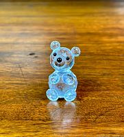 Front view of clear faceted crystal teddy bear figurine with black eyes and nose, placed on wooden surface, detailed facets visible.