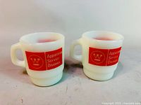 Two white opaque milk glass mugs with red Apparatus Service Division logo and handles, shown side by side on a neutral surface.