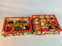 Two original red cardboard boxes with green holly and white snowflake holiday graphics visible. Each box has compartments containing vintage glass Christmas ball ornaments in red, green, gold, white, and multicolor tones.