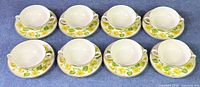 Seven bouillon cups each paired with a matching saucer, arranged on a surface showing the floral yellow and green leaf pattern.