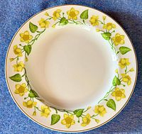 Top view of the round fine bone china chop plate with yellow Kingcup flowers and green leaves on white background with gold rim.