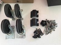 Four black rubber caster wheels with metal mounting plates displayed lying on a table, accompanied by various small metal and plastic hardware pieces including screws and mounting brackets.