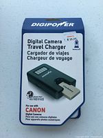 Front of packaging showing Digipower branding, product name Digital Camera Travel Charger, compatible with Canon cameras, and key features like LCD display and USB port.