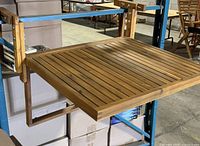 Foldable wooden balcony table mounted on a metal shelf, showing slatted table top and foldable arm supports, with natural wood color and grain visible.